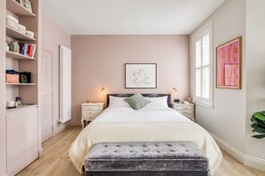 A bedroom with a bed, bedside tables, a nightstand, a bench, a radiator, a shelf with books and decorative items, and a framed picture on the wall.