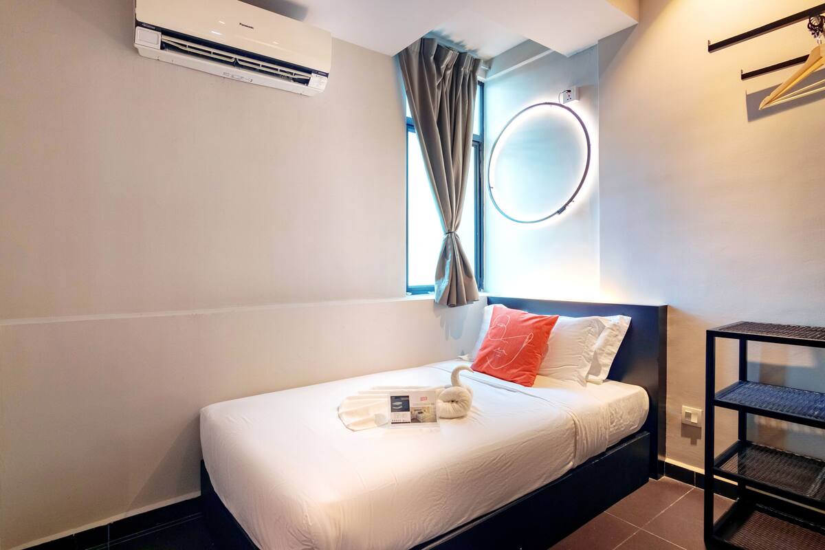 A neatly made bed with a white comforter and a swan-shaped pillow. There is a small table with a tray of toiletries and a towel. A wall-mounted air conditioner is visible, and a window with curtains is present.