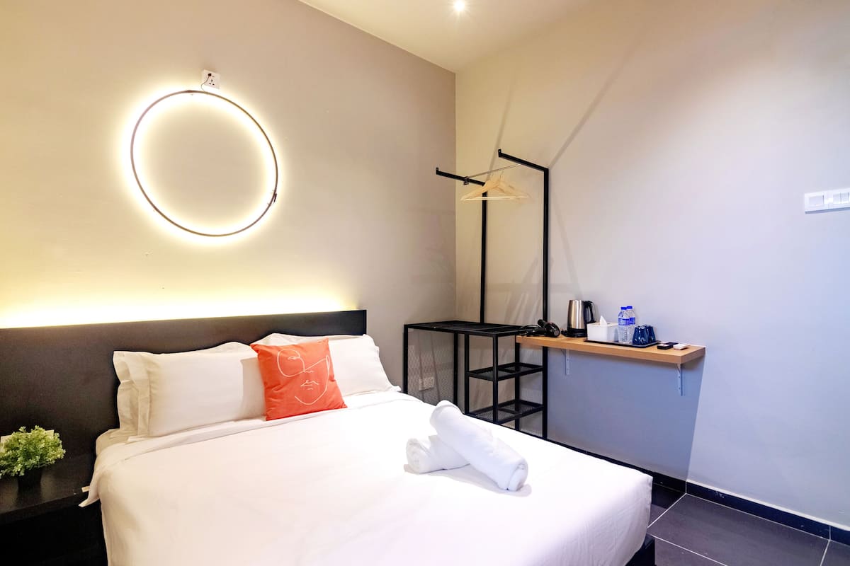 A hotel room with a neatly made bed, a bedside table with a kettle and a bottle, a hanging light fixture, and a small shelf with toiletries.