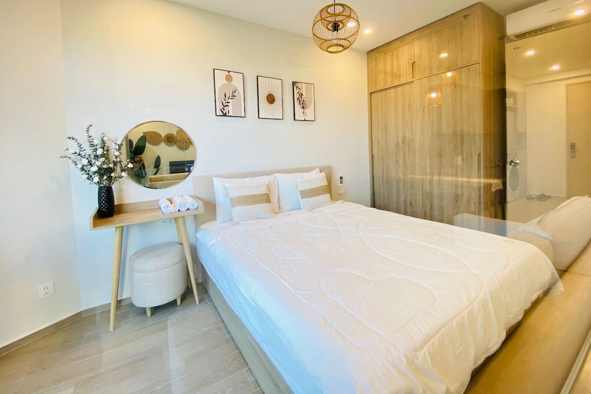A modern bedroom with a large bed, a wooden wardrobe, a desk with a mirror, and a vase with flowers.