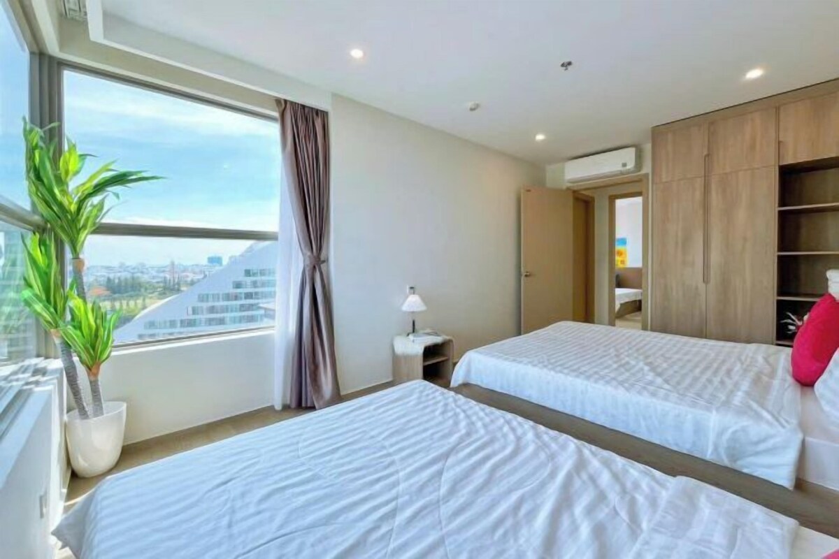 A hotel room with two beds, a large window with a city view, a potted plant, and a wardrobe.