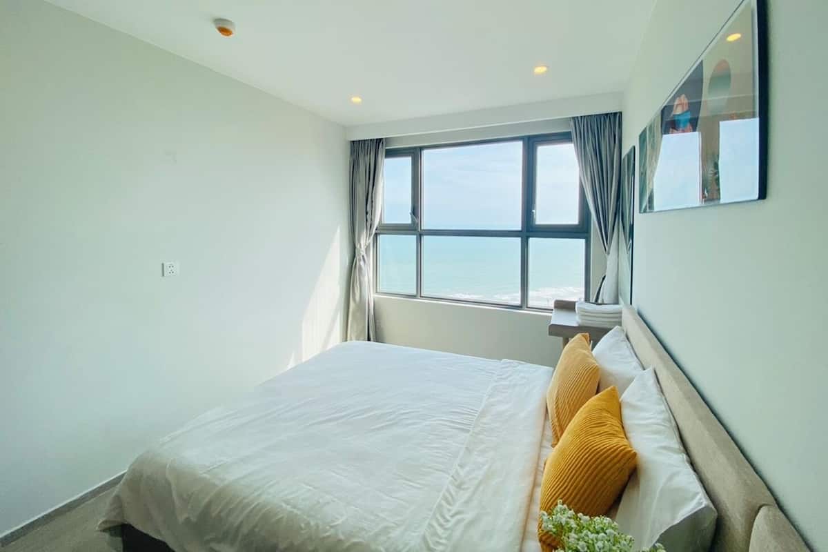 A bedroom with a large bed, a window with curtains, and a view of the sea.