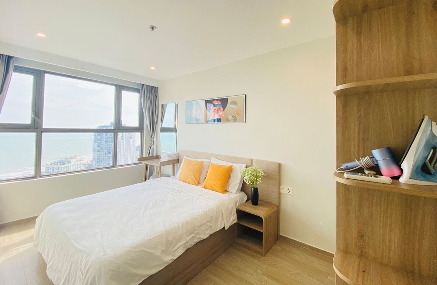 A modern hotel room with a large bed, a wooden nightstand, a small table, and a view of the city through large windows.