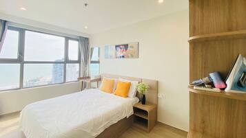 A modern hotel room with a large bed, a wooden nightstand, a small table, and a view of the city through large windows.