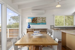 Uma sala de jantar com uma mesa de madeira, cadeiras brancas, um abajur azul e um quadro na parede.