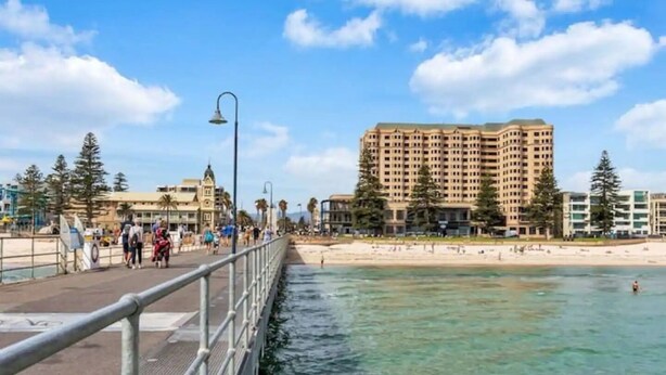 A coastal promenade with a beach, a pedestrian walkway, and a large hotel building in the background. - Sunsets at Glenelg - entire 3 bedroom apartment
 (Glenelg)