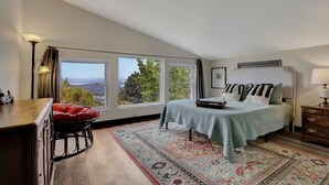 Un dormitorio con un ventanal grande, una cama con colcha verde, una silla roja, un aparador de madera y una alfombra estampada.