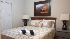 A neatly made bed with a patterned bedspread, two pillows, and two towels. There is a painting on the wall above the bed, and two bedside lamps on either side. - Clean Comfy Cottage - Near Downtown High Point Furniture Market, Hospitals & HPU (High Point)