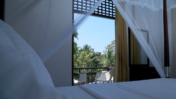 Un dormitorio con cama, cortinas blancas y vistas a palmeras y un balcón con sillas blancas.
