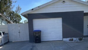 A garage with a white door, a black trash bin, and a concrete driveway. - COMFORTABLE ORLANDO PRIVATE STUDIO W/FULL KITCHEN! (Orlando)