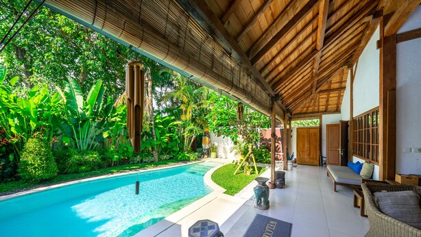 Ein Poolbereich mit einer Holzpergola, üppiger Bepflanzung und einer Sitzgelegenheit mit einer Bank und Korbmöbeln.