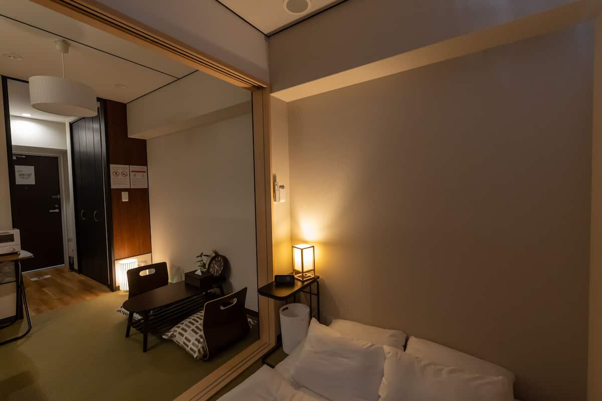 A hotel room with a bed, a desk with a chair, a lamp, and a mirror.