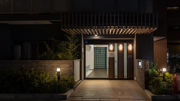 A modern building entrance with a wooden overhang, a pathway leading to the door, and a well-lit facade. - H2H Tsutenkaku (Osaka)