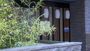 A bamboo plant in a stone planter outside a building with a wooden door and a sign on the wall. - H2H Tsutenkaku (Osaka)