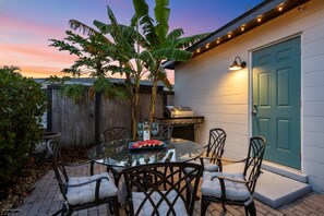 Une terrasse avec une table ronde en verre, quatre chaises, un barbecue et une porte bleue.