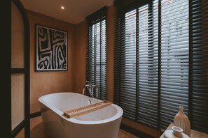 Een moderne badkamer met een vrijstaand bad, een groot abstract schilderij en een raam met zonwering.