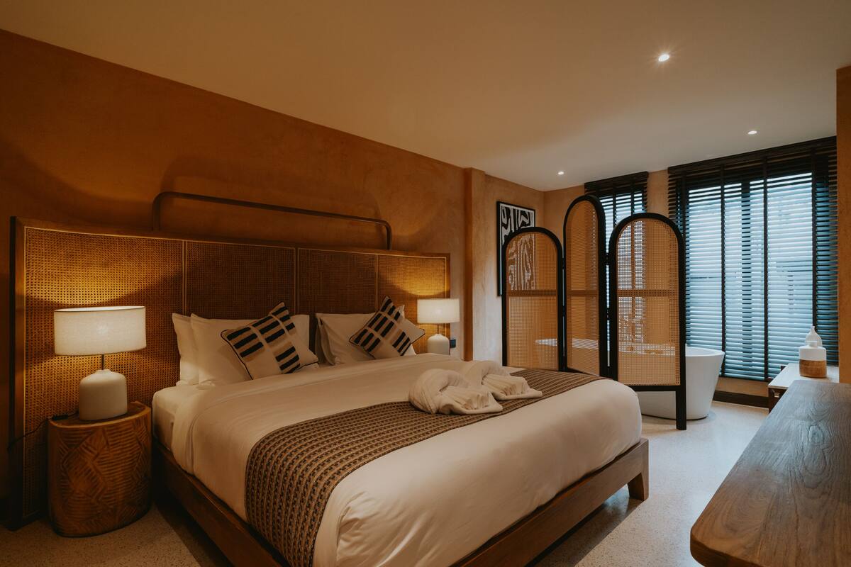 A hotel room with a large bed, two bedside lamps, a wooden headboard, a rattan chair, a wooden table, and a window with blinds.