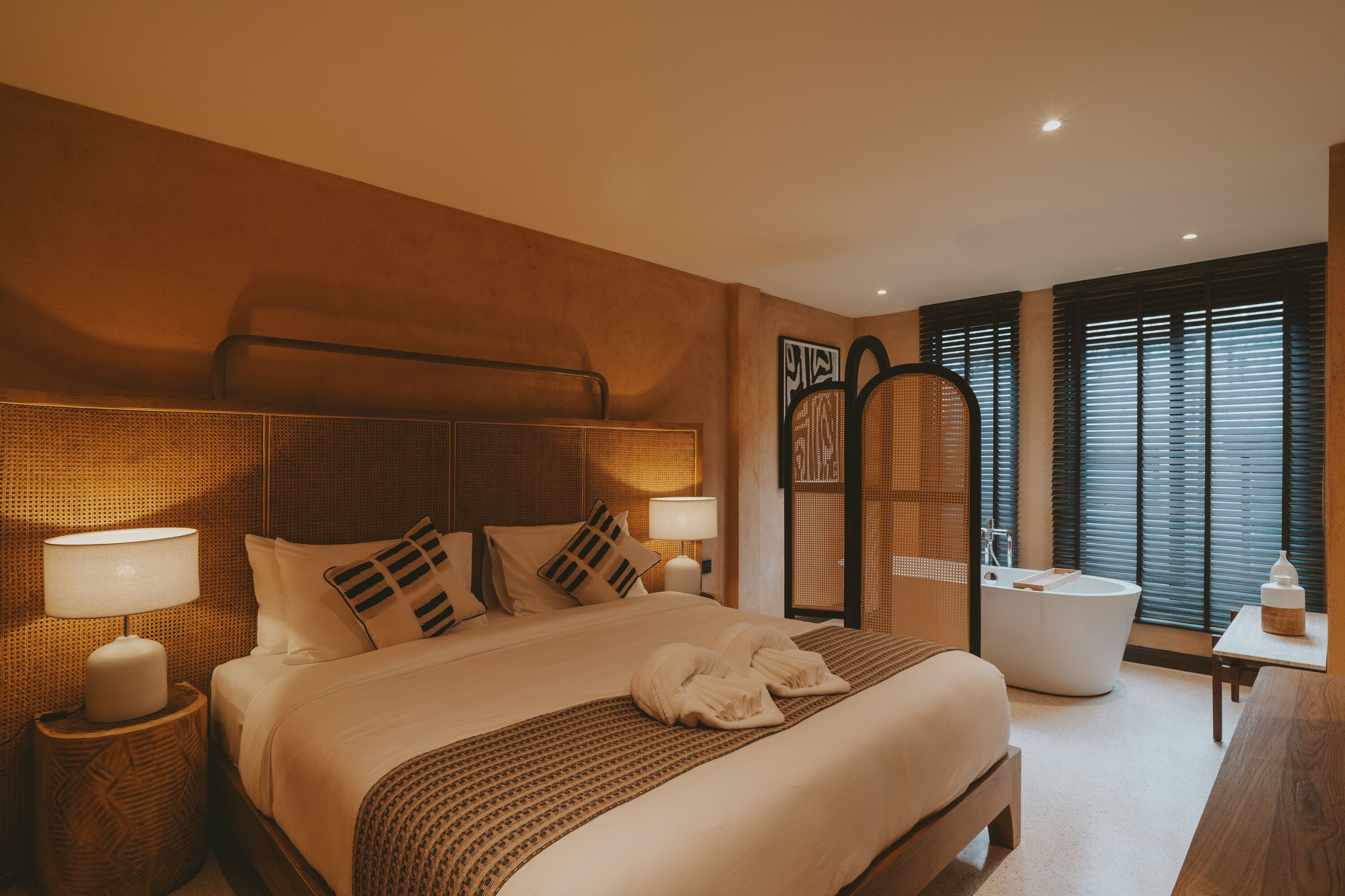 Ett modernt hotellrum med en stor säng, en sittgrupp med en fåtölj och ett bord, ett badkar och ett stort fönster med persienner.