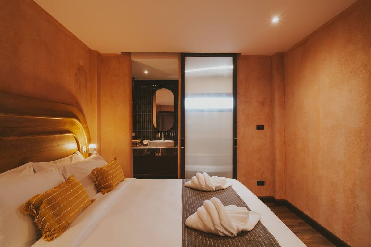 A hotel room with a wooden headboard, a bed with white linens, a bedside table with a lamp, and a bathroom visible through an open door.