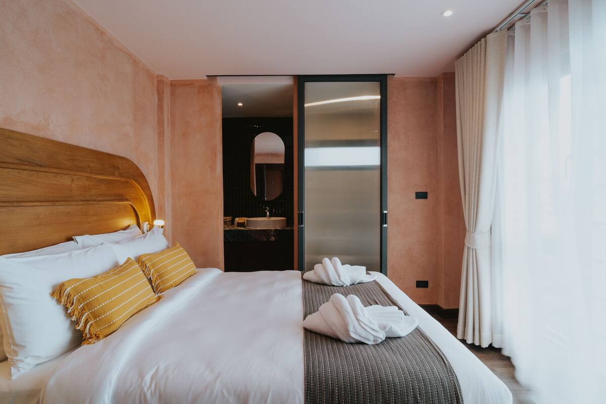 A hotel room with a large bed, a wooden headboard, a bathroom with a mirror and sink, and a sliding door to a balcony.