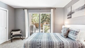 Quarto com cama grande, uma cadeira, varanda com vista para árvores e uma porta.