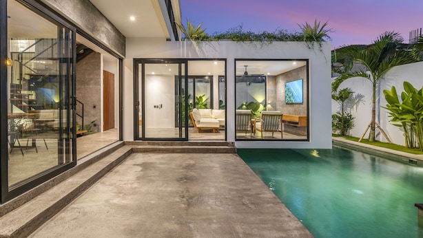 Una casa moderna con un ventanal de vidrio que da a una sala de estar, una piscina y un comedor con mobiliario exterior.