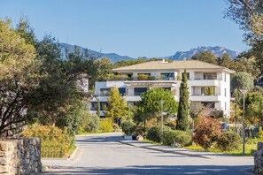 Un immeuble résidentiel moderne à plusieurs étages, doté de balcons, entouré d’une végétation luxuriante et avec des montagnes en toile de fond.