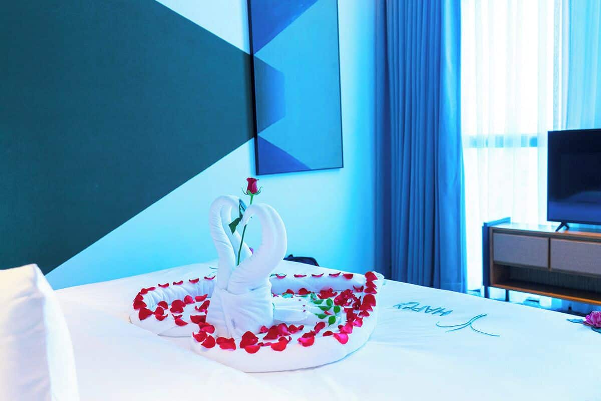 Una habitación de hotel con una cama decorada con pétalos de rosa y un centro de mesa con forma de cisne.