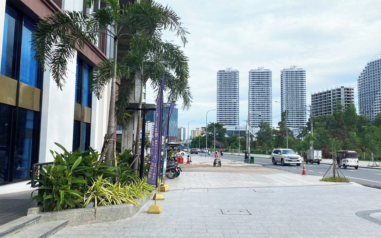 A modern urban street with tall buildings, a sidewalk lined with palm trees, and a few pedestrians.