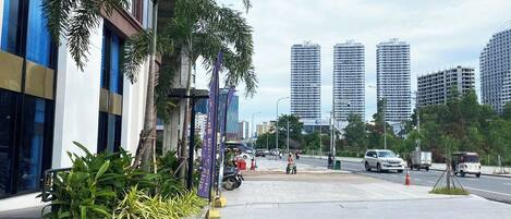 A modern urban street with tall buildings, a sidewalk lined with palm trees, and a few pedestrians.