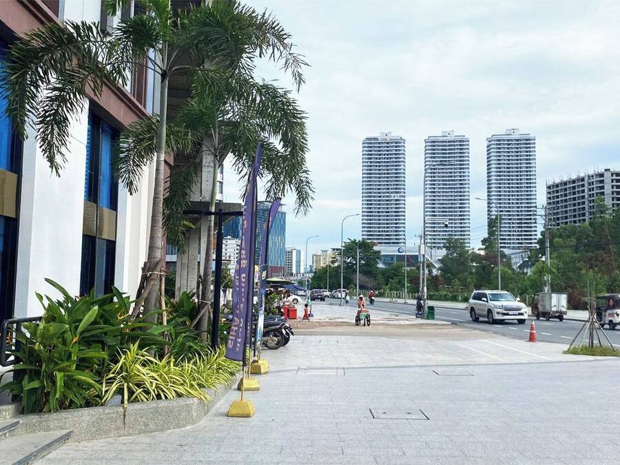 A modern urban street with tall buildings, a sidewalk lined with palm trees, and a few pedestrians.
