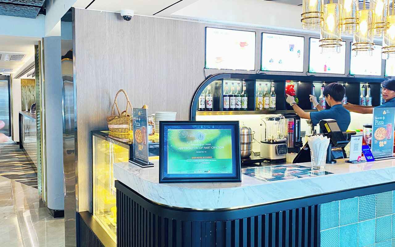 A hotel reception area with a digital display, a counter with a menu, and a bar area with staff.