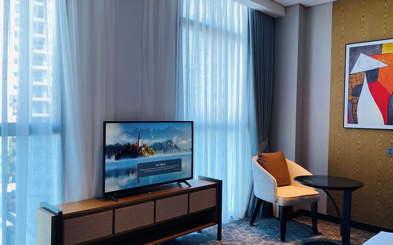Una habitación de hotel moderna con una televisión de pantalla plana grande sobre un mueble de madera, un sillón cómodo, una pequeña mesa redonda y un ventanal con cortinas ligeras.