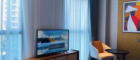 Una habitación de hotel moderna con una televisión de pantalla plana grande sobre un mueble de madera, un sillón cómodo, una pequeña mesa redonda y un ventanal con cortinas ligeras.
