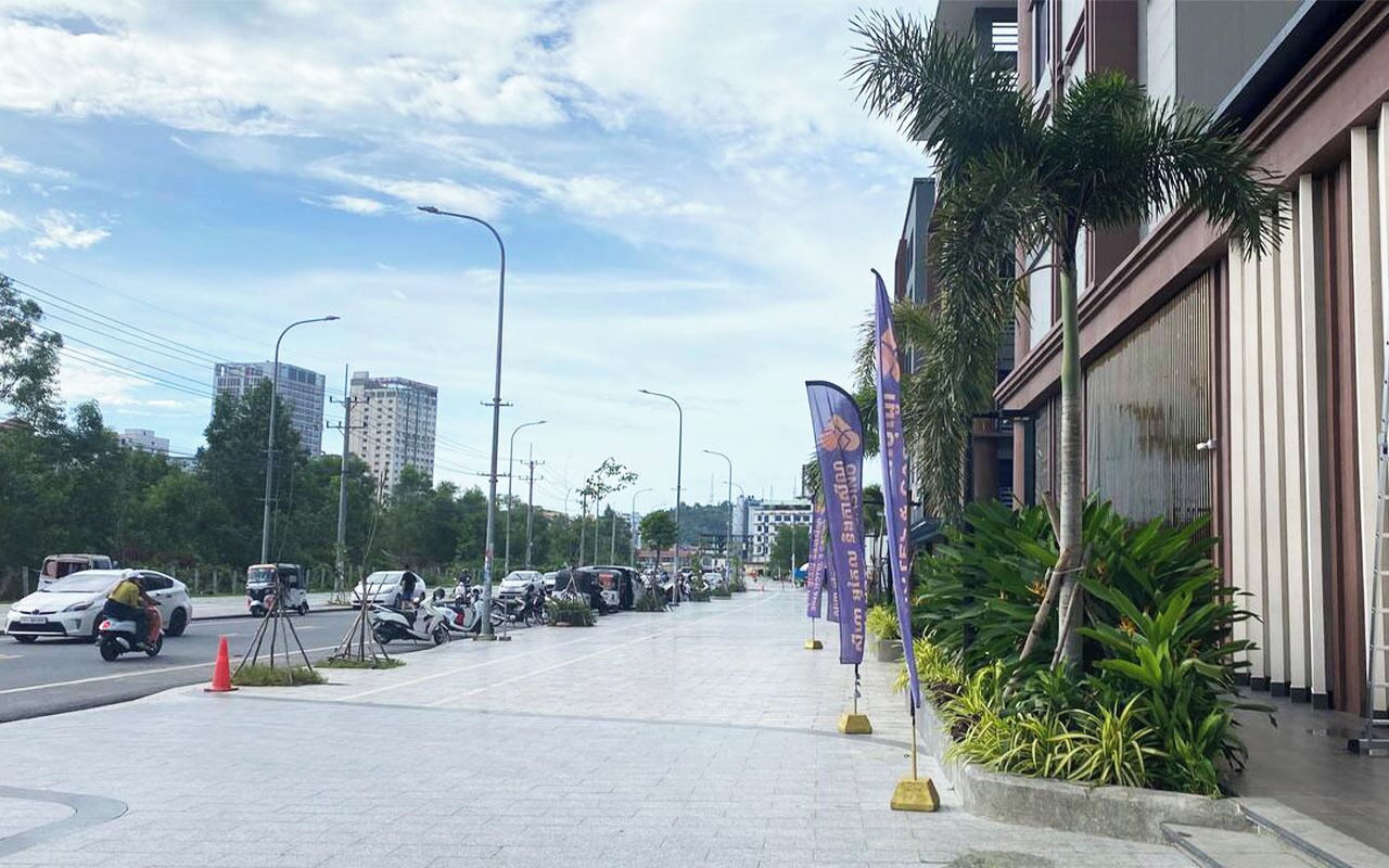 A street with parked cars and motorcycles, a sidewalk, and buildings with flags.