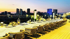 A rooftop dining area with tables set for a meal, wicker chairs, and a city skyline view in the background.