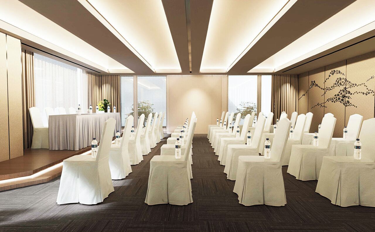 Un salón de conferencias con filas de sillas y mesas blancas, amplias ventanas y una zona de escenario.