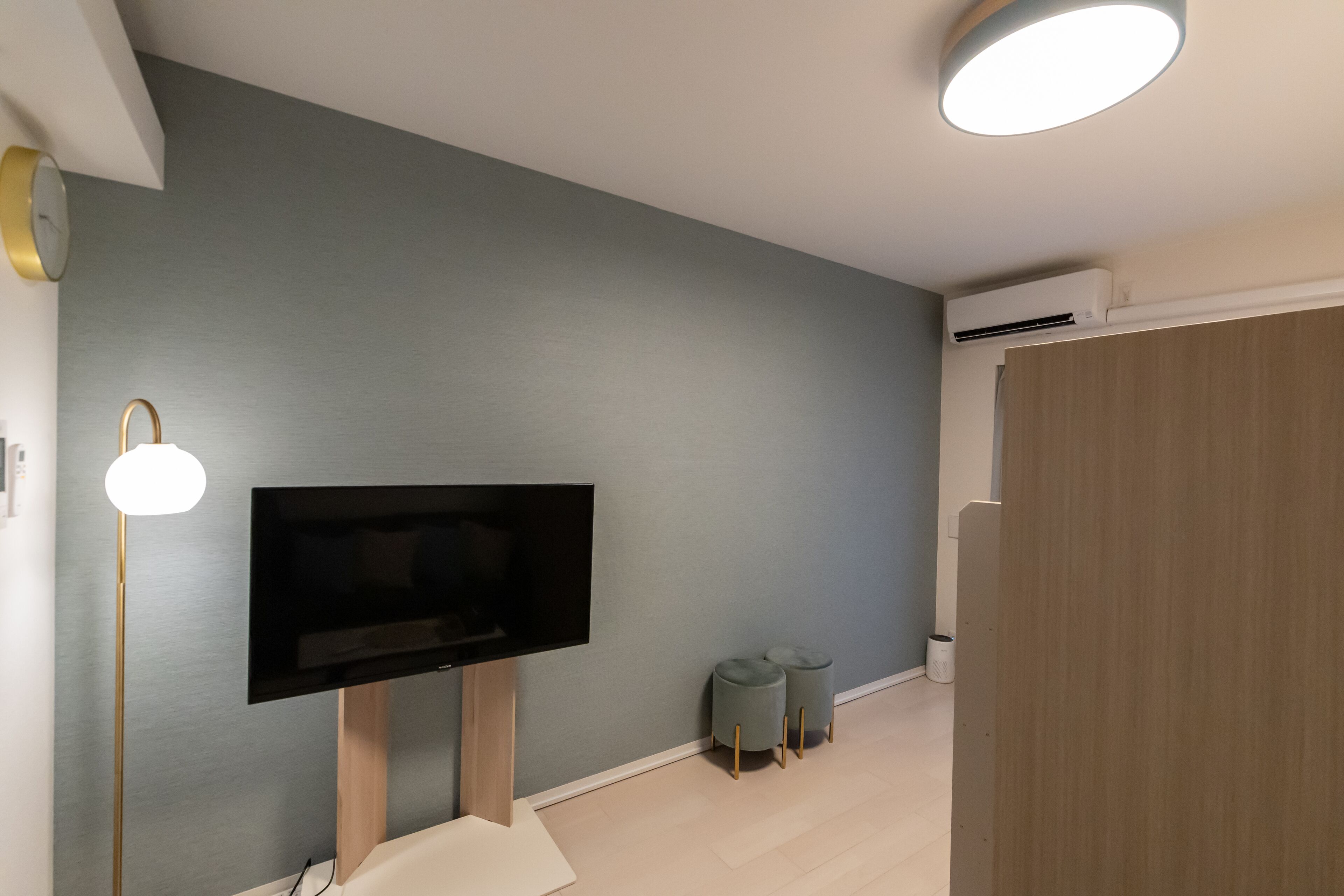 Ein modernes Wohnzimmer mit einem an einer Holzkonstruktion befestigten Flachbildfernseher, einer Stehlampe und zwei grauen Hocker.