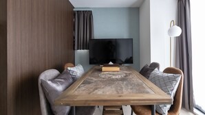 A modern living room with a wooden dining table, grey upholstered chairs, a flat-screen TV, and a floor lamp.