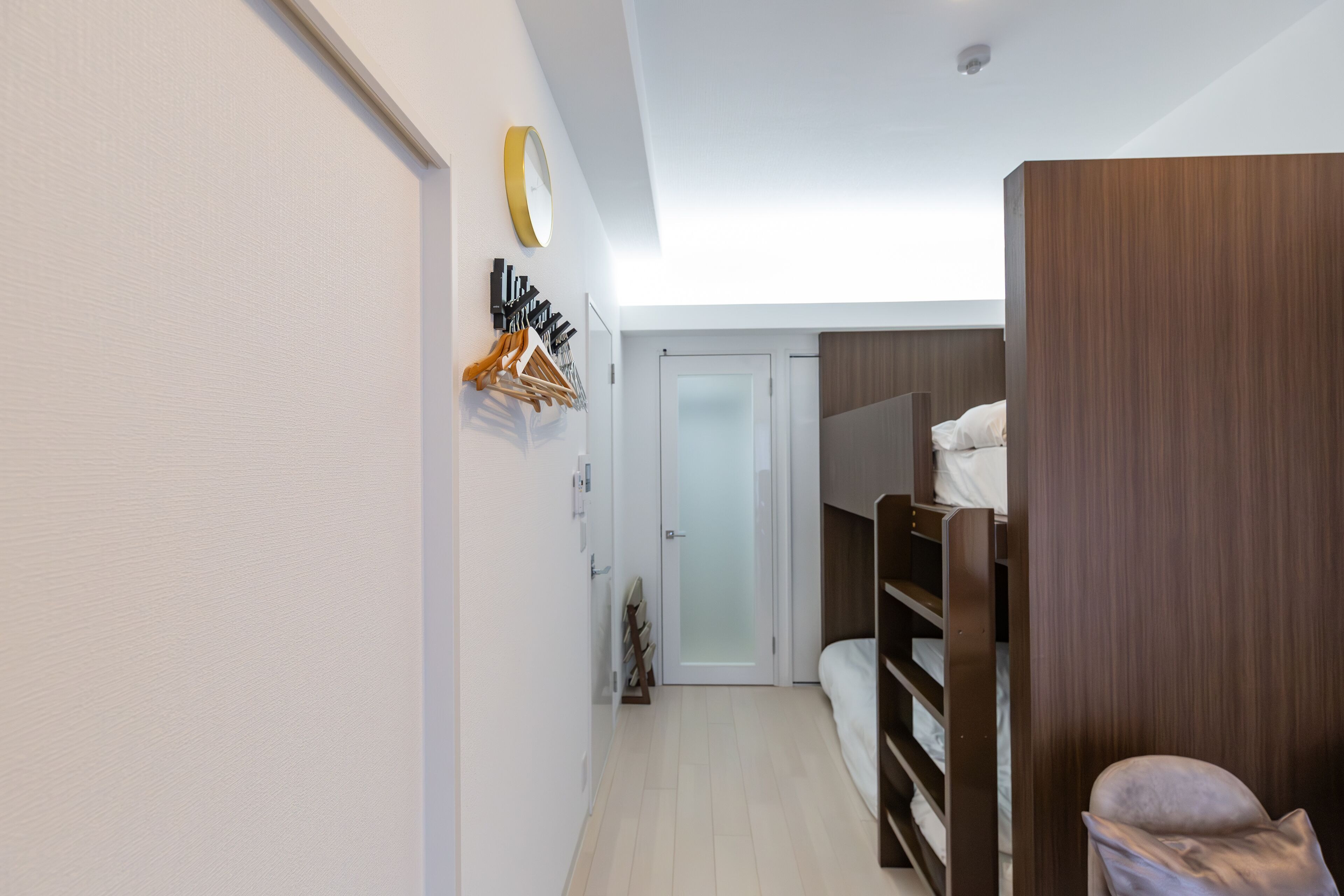 A narrow hallway with a wooden wardrobe, a bed with a white mattress, and a coat rack with hanging items.