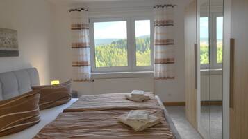 Ein Schlafzimmer mit einem Bett, zwei Kissen, einem Fenster mit Vorhängen, einem Heizkörper und Blick auf Bäume.