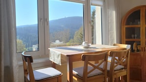 A dining area with a table and chairs, large windows with a view of mountains and trees, and a wooden cabinet. - Oberwiesenthal, Unweit der Skipiste (Oberwiesenthal)