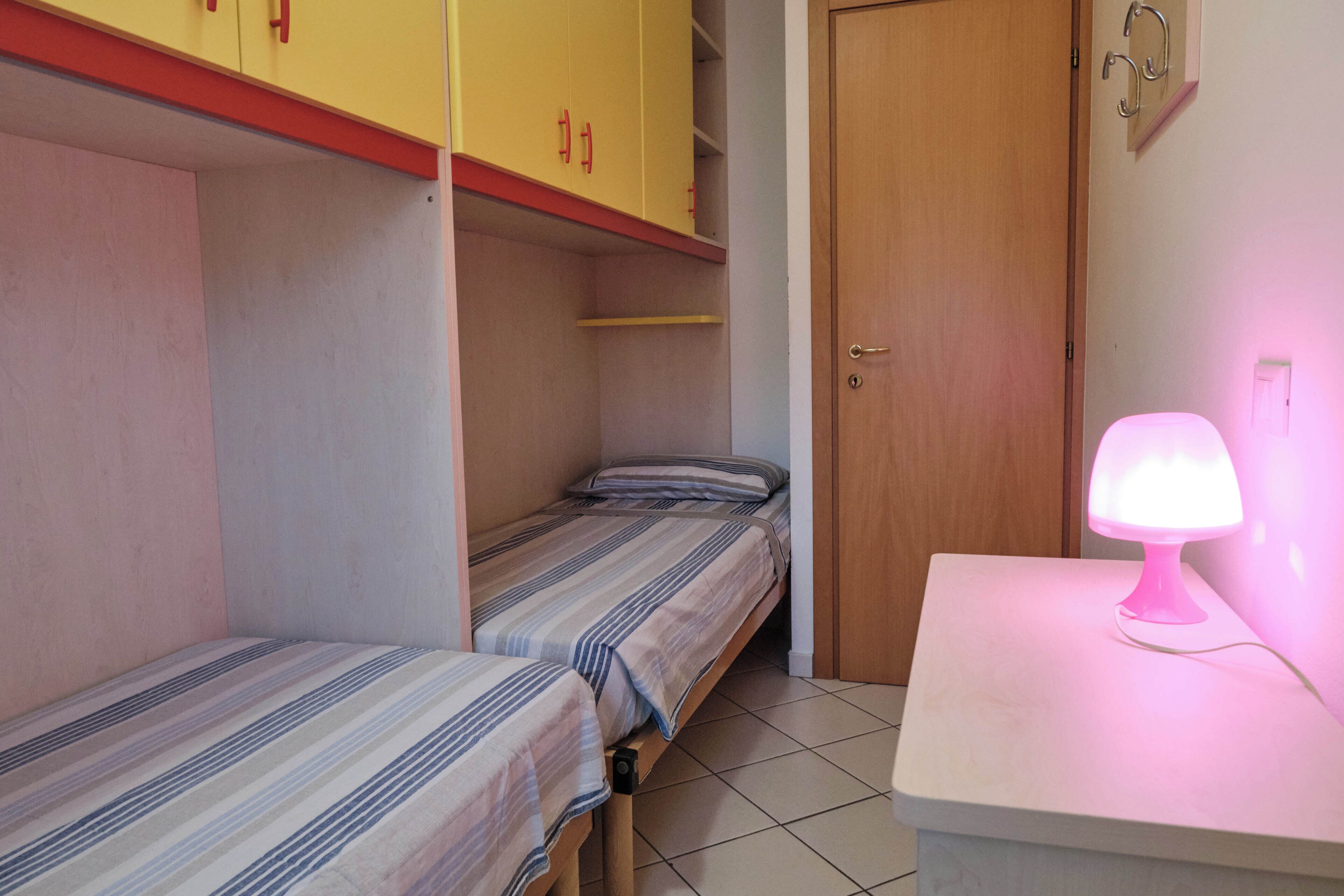 A small room with two single beds, a wooden door, and a bedside table with a pink lamp.