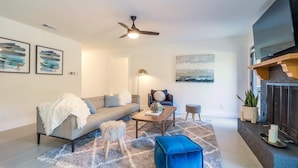 A modern living room with a sofa, coffee table, and ottoman, a fireplace, and a ceiling fan. - ATX Oasis retreat - pool, hot tub, games & more! Trendy modern setup. (Austin)