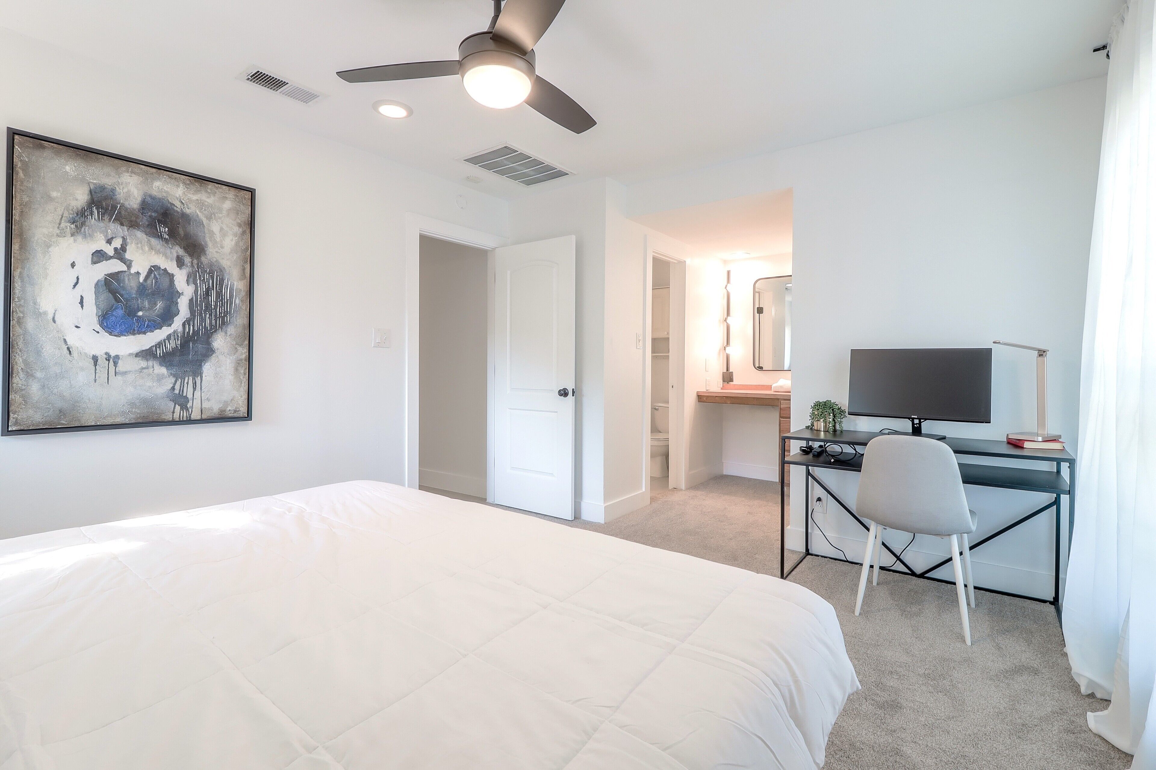 A bedroom with a bed, a desk with a chair, a ceiling fan, and a large abstract painting on the wall.