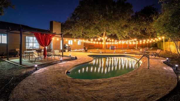 A pool area with a curved edge, surrounded by a sandy deck, and string lights overhead. - ATX Oasis retreat - pool, hot tub, games & more! Trendy modern setup. (Austin)