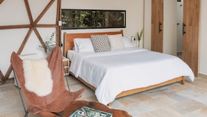 A bedroom with a wooden bed, a brown leather chair, a wooden coffee table, and a large window with a view of trees. - TERRAMIA ESTATE (Santa Marta)