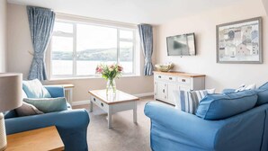 A living room with a blue sofa, a small table with a vase of flowers, a TV on the wall, and a view of the outdoors through large windows. - High Tide -  a house that sleeps 6 guests  in 3 bedrooms (Salcombe)