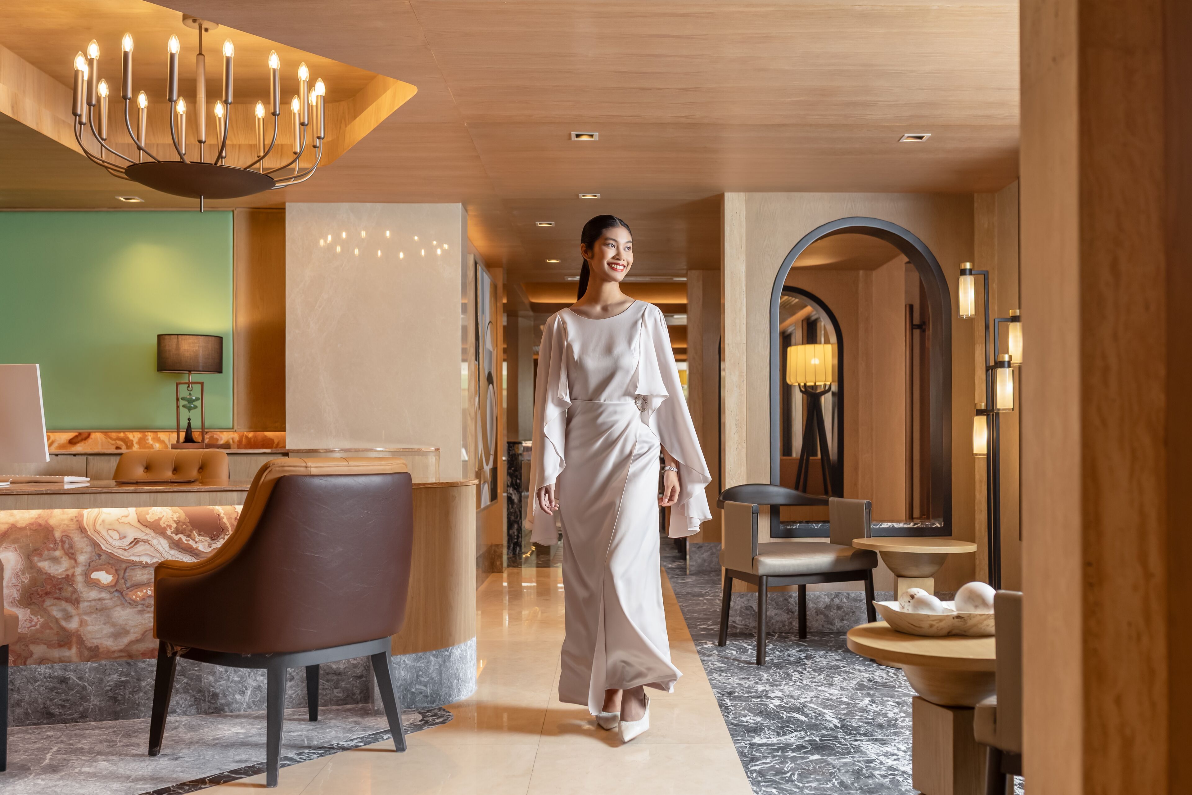 A modern hotel lobby with a marble reception desk, a seating area, and a large chandelier.