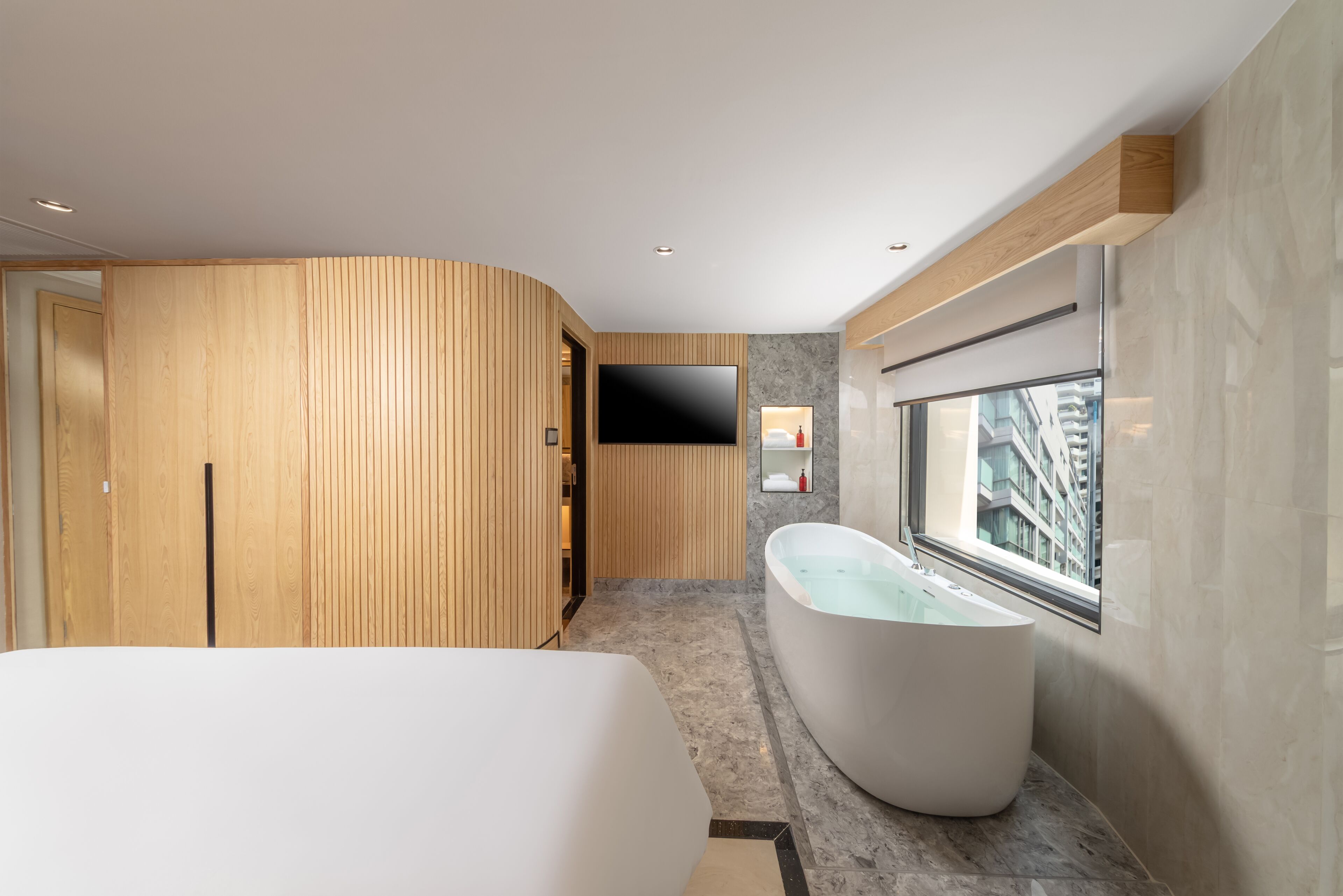 A modern bathroom with a freestanding bathtub, wooden paneling, a flat-screen TV, and a large window offering a city view.